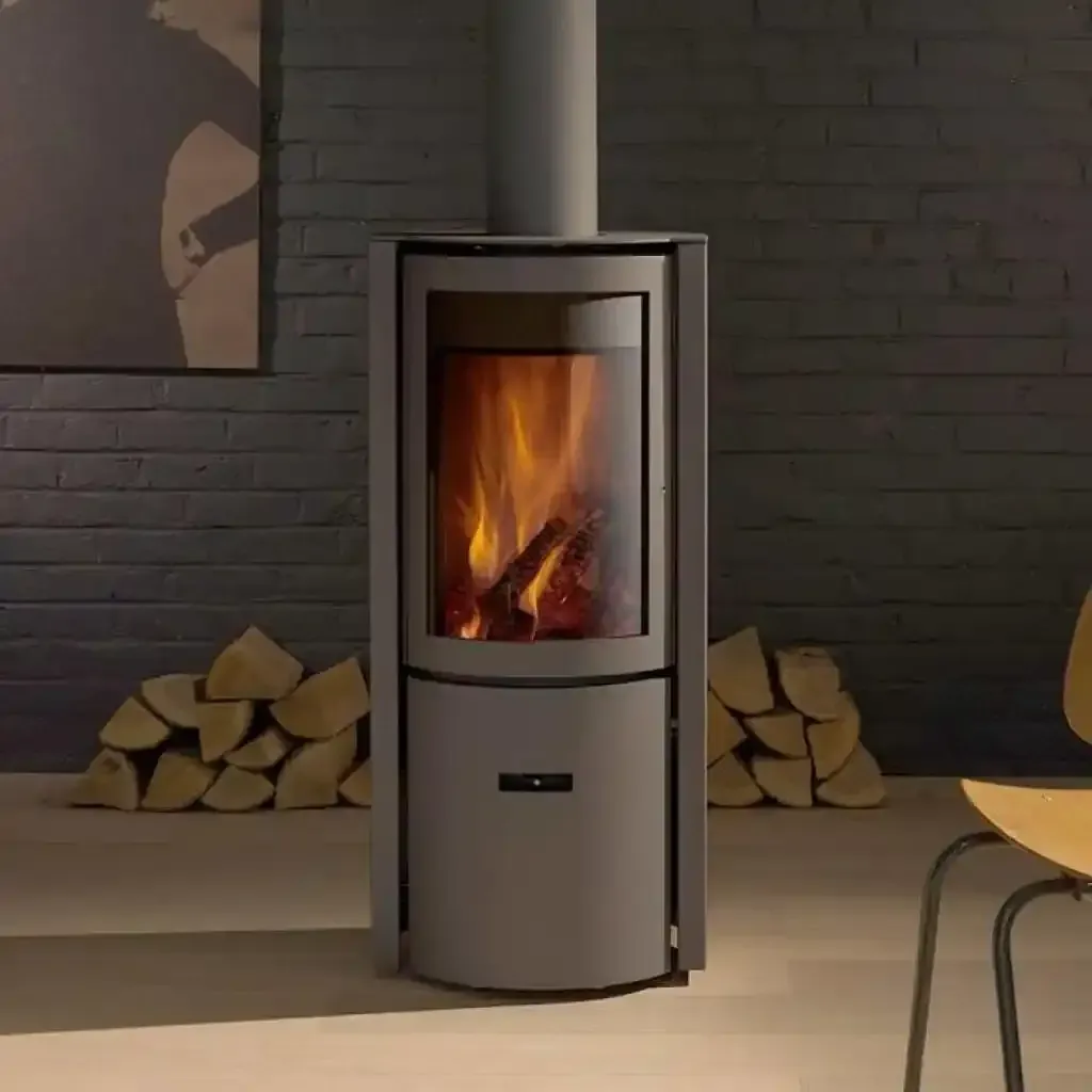 Modern gray wood-burning stove with flames in a room with brick wall, firewood, and a chair.