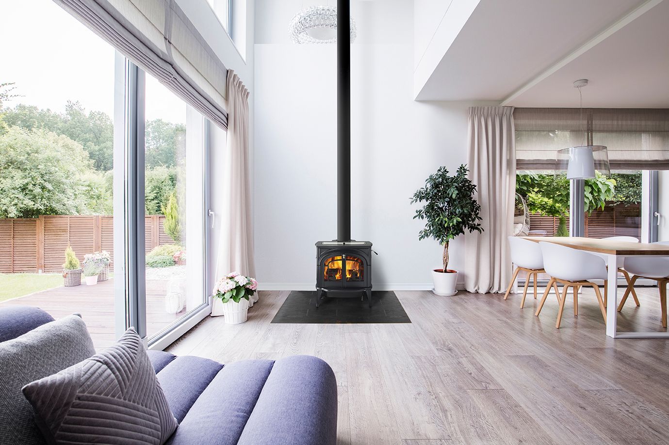 Bright open living room with a modern fireplace, gray sofa, dining area, and large windows overlooking greenery