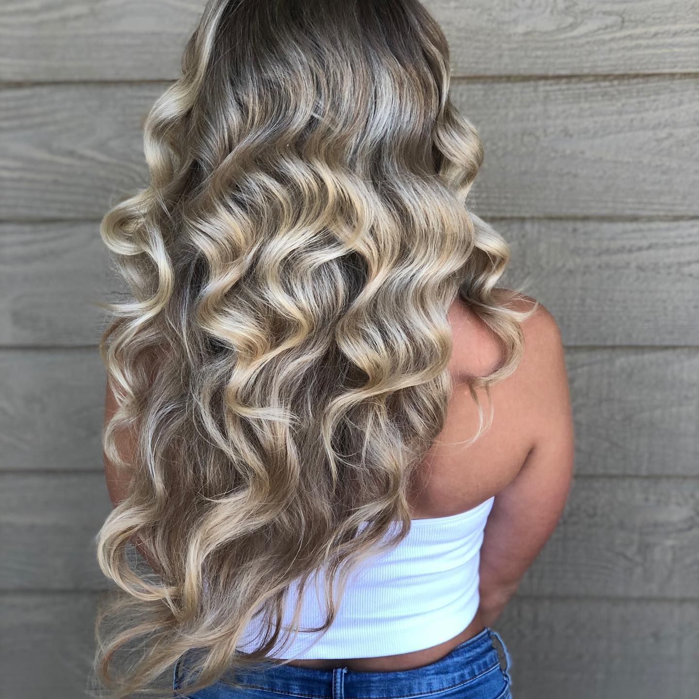 A woman with long blonde curly hair is standing in front of a wooden wall.