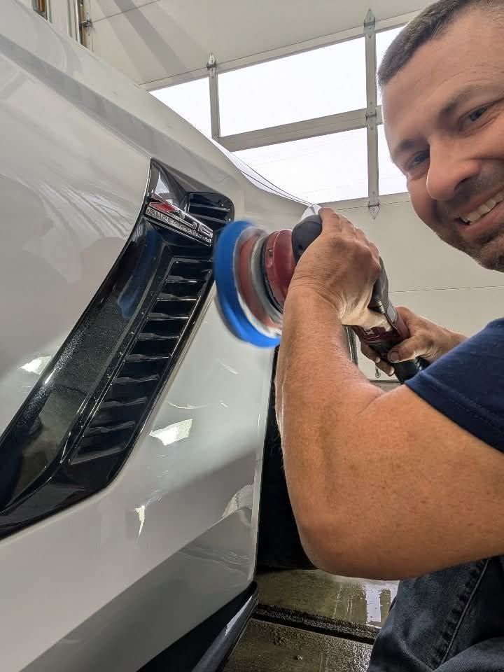 Man polishing a white car panel with a buffer in a garage, smiling at the camera