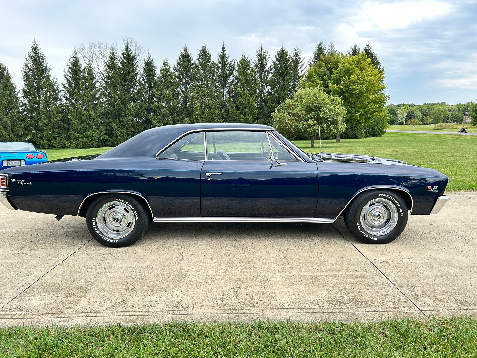 Dark blue classic car parked on a concrete lot with trees and grass in the background