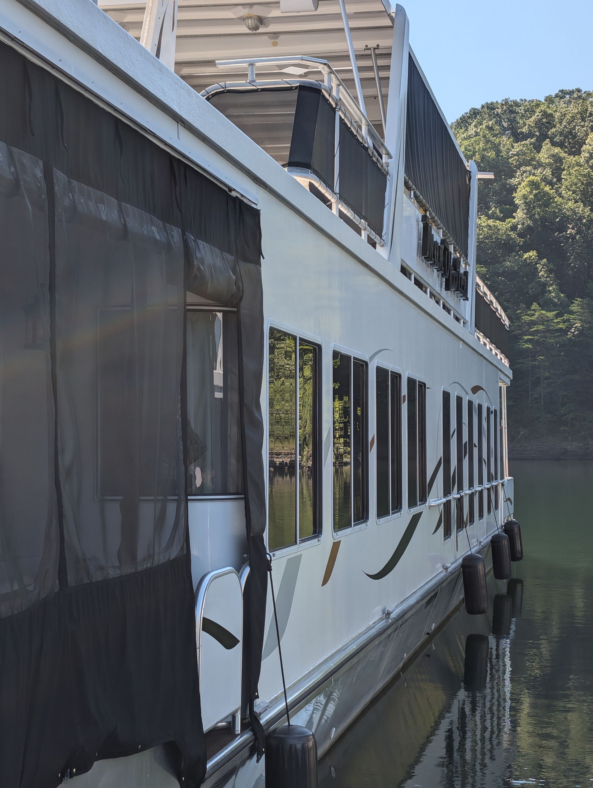 White houseboat docked beside a tree-lined river, with large windows and a covered deck.