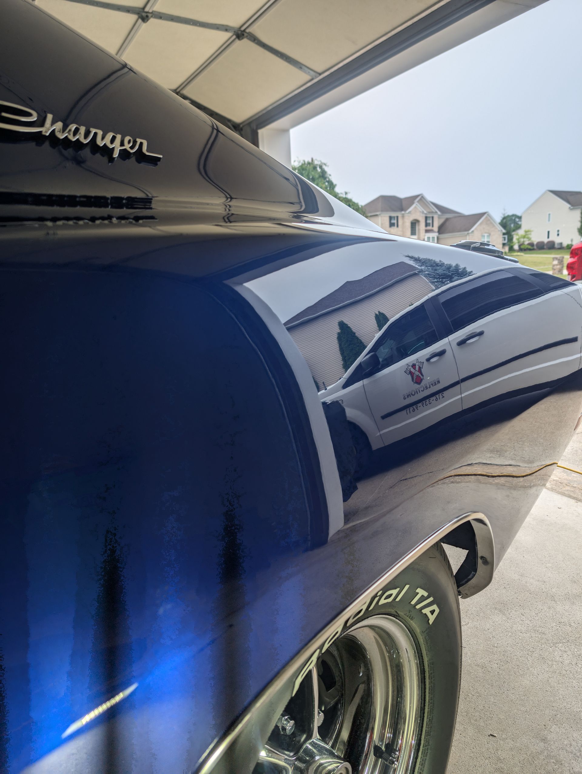 Blue classic car rear quarter in a driveway, with chrome wheel and residential houses in the background.