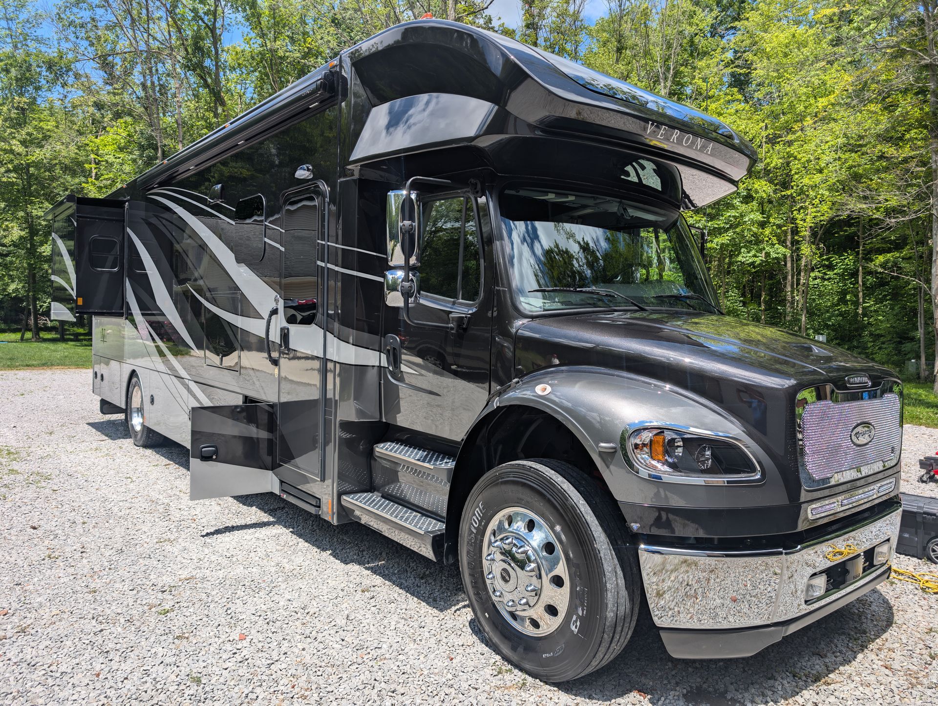 Black and silver luxury motorhome parked on a gravel lot beside trees