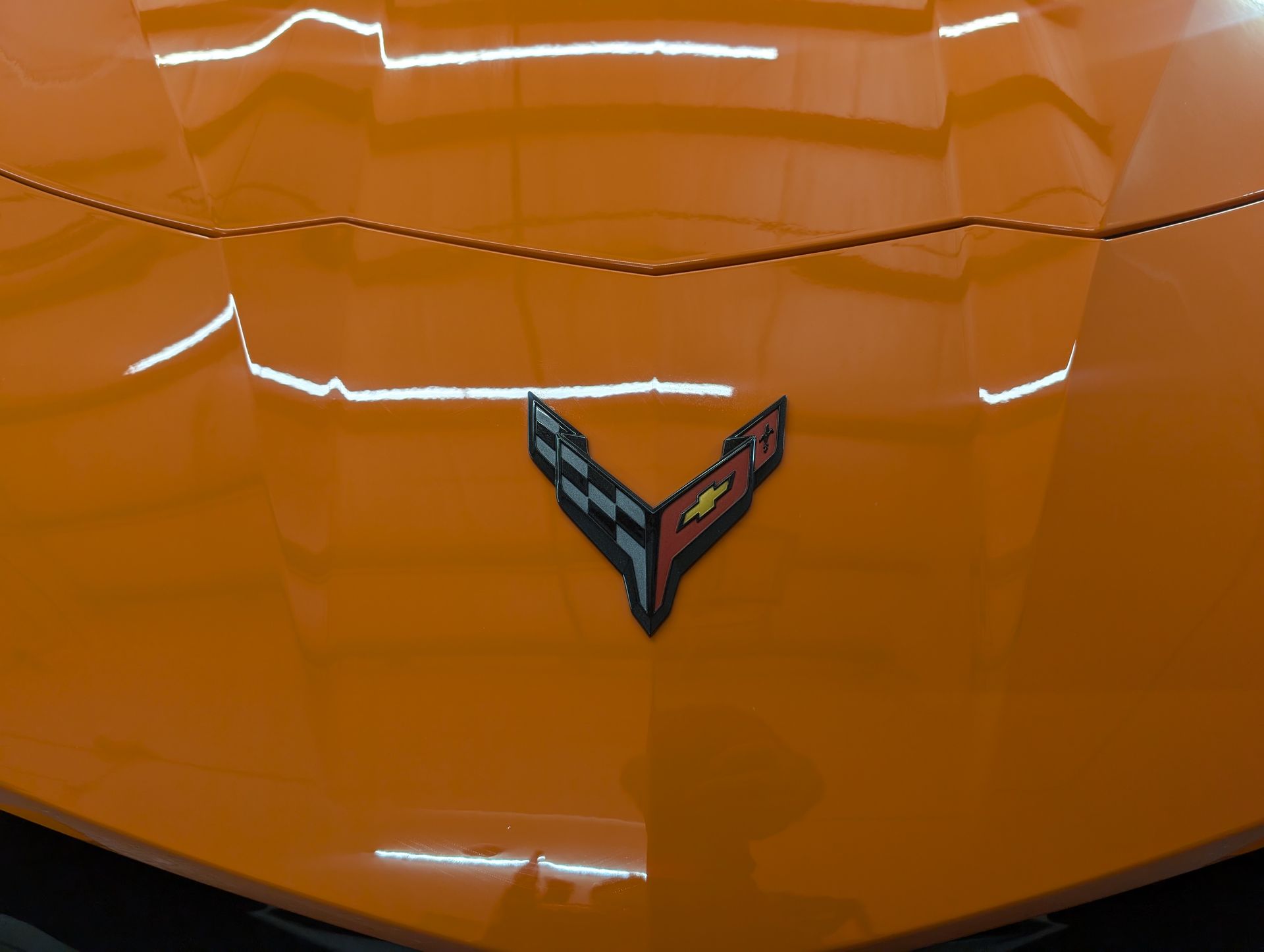 Close-up of an orange car hood with a Corvette emblem centered beneath reflections.