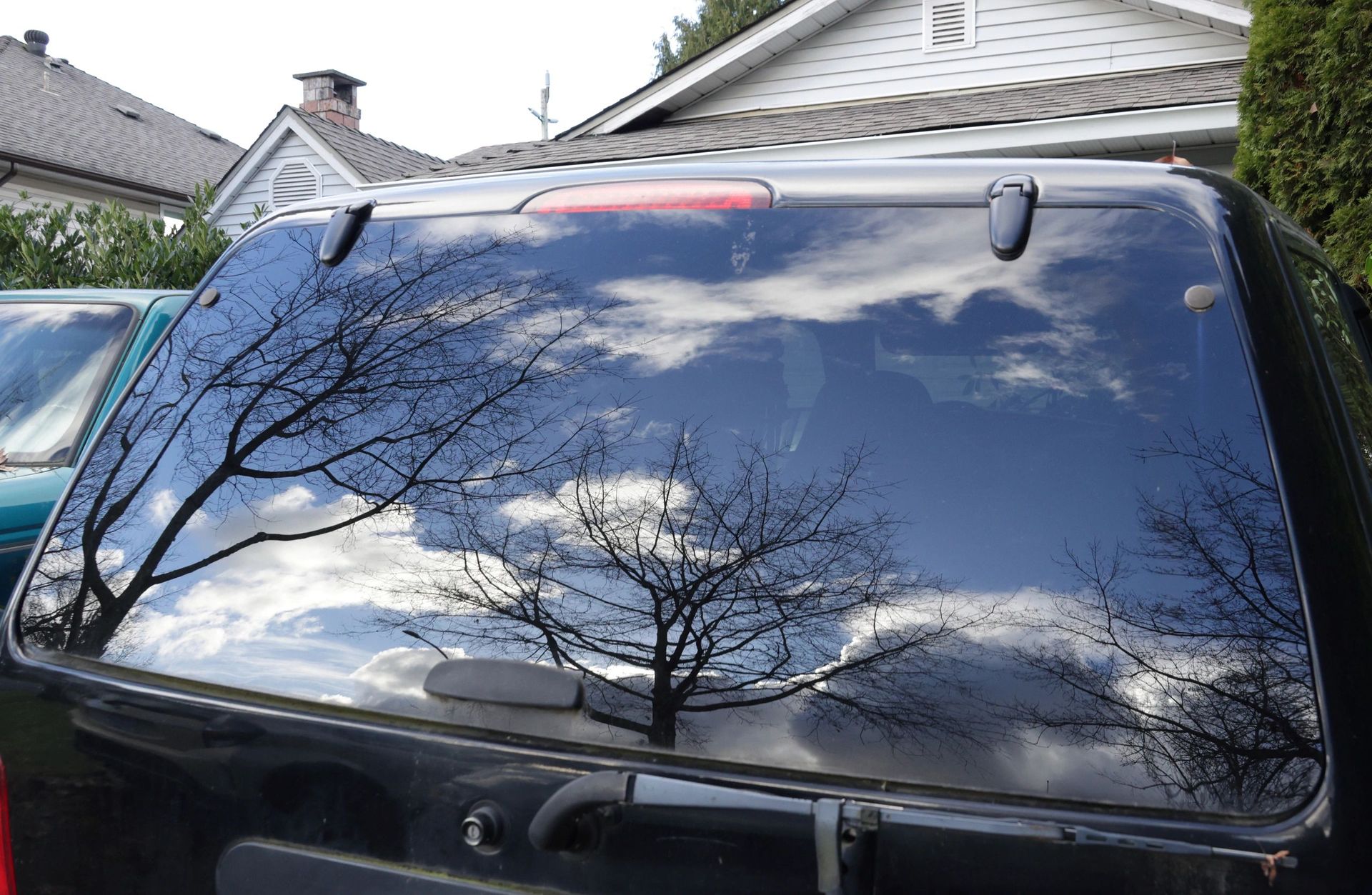 Rear window of a black SUV reflecting bare trees and clouds in a suburban driveway