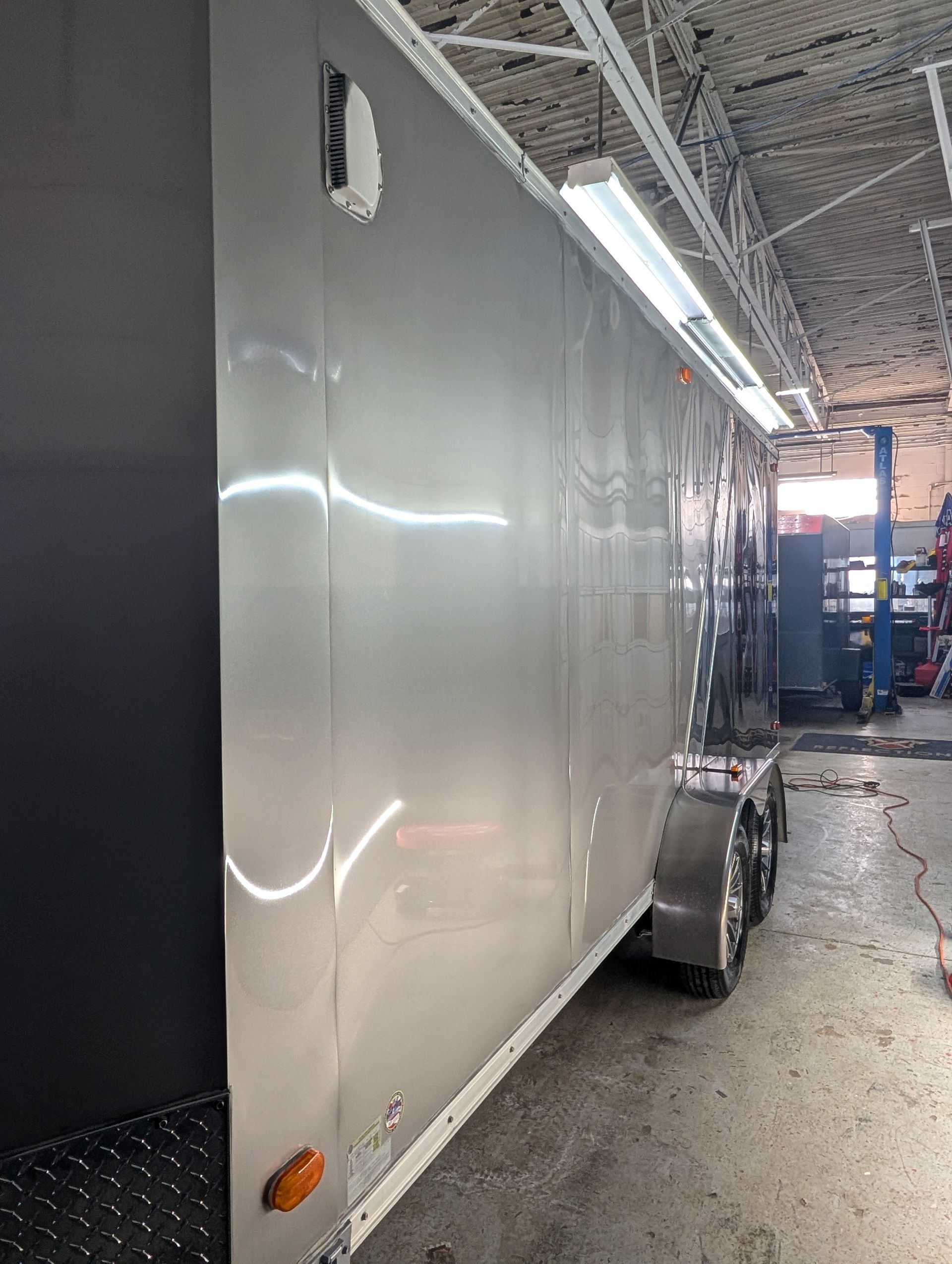 White enclosed trailer inside a workshop garage, with a rear wheel visible and overhead lights reflected on its side.