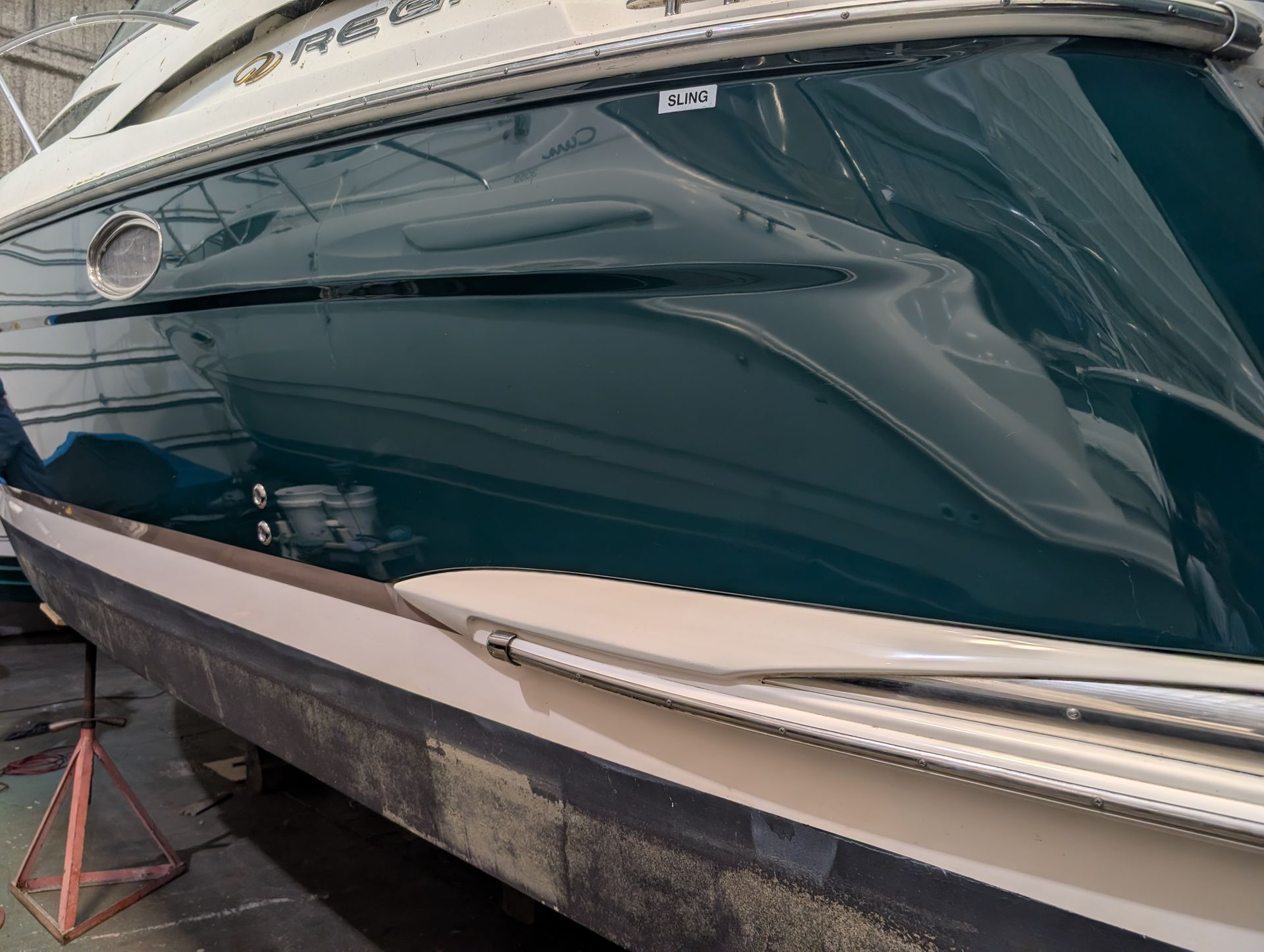 Close-up of a dark teal yacht hull with reflections, docked beside a dock in a boatyard.