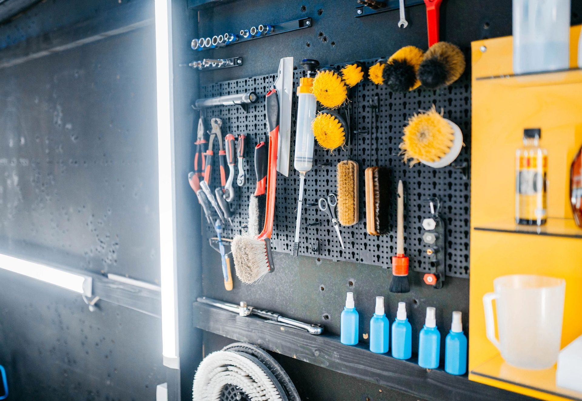 Tool wall in a workshop with hanging brushes, wheels, bottles, and yellow storage cabinets.