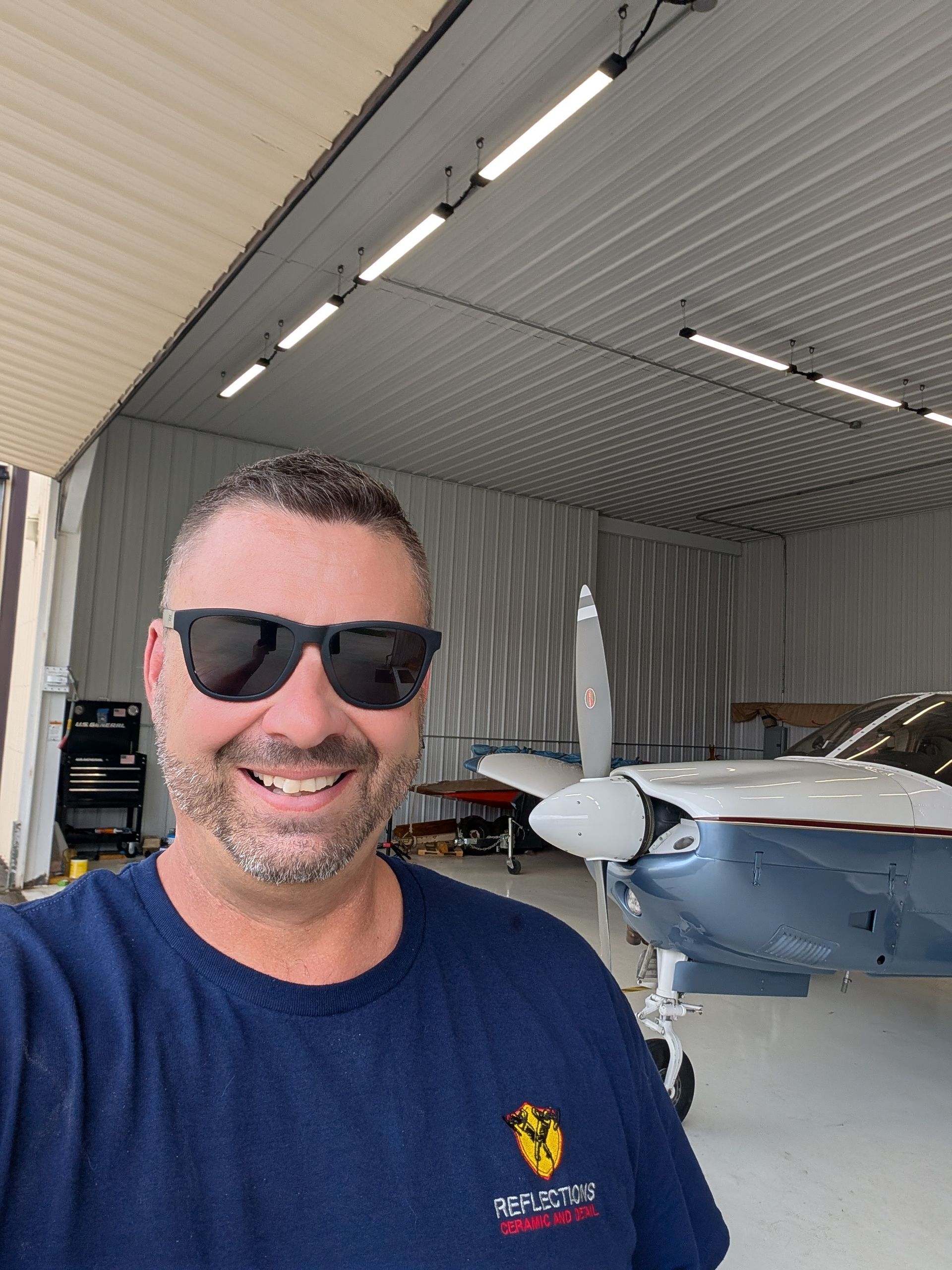 Smiling man in sunglasses standing in a hangar beside a small airplane
