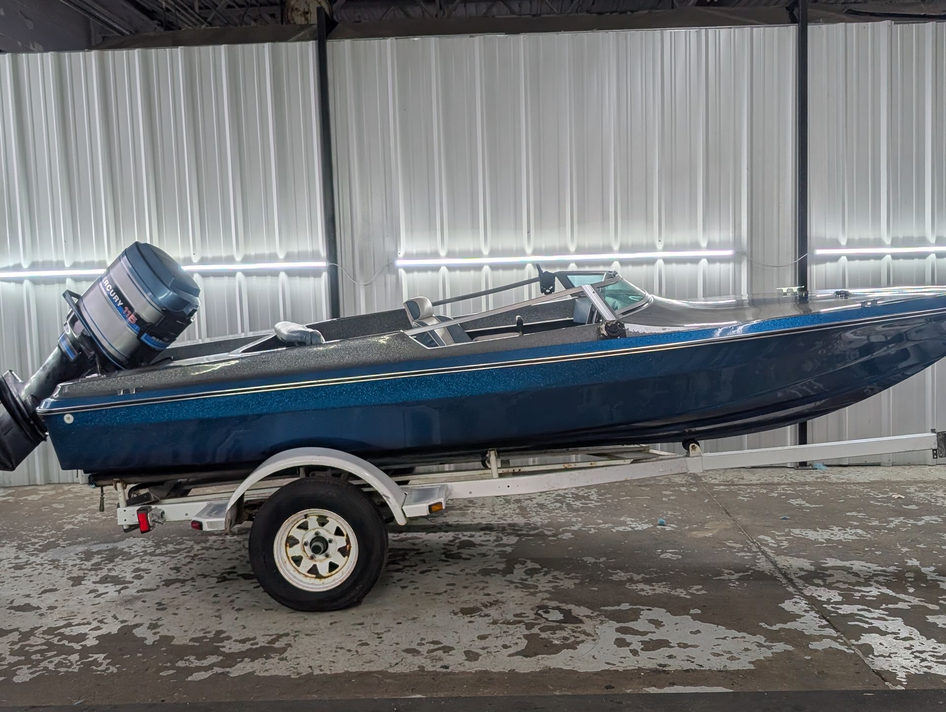 Blue fishing boat on a trailer in an indoor warehouse, with an outboard motor attached