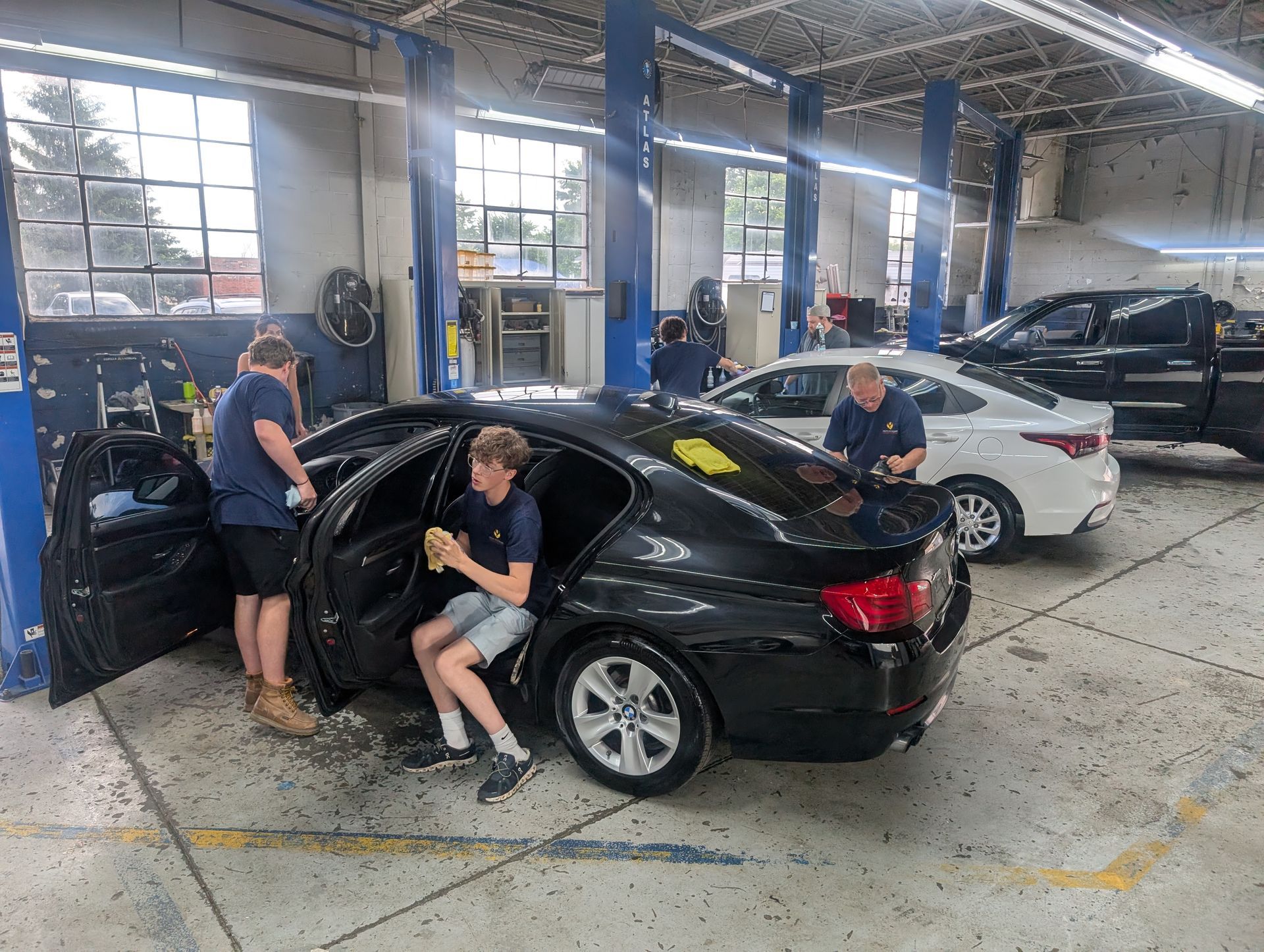 Three mechanics clean a black sedan in an auto repair shop with other cars in bays.