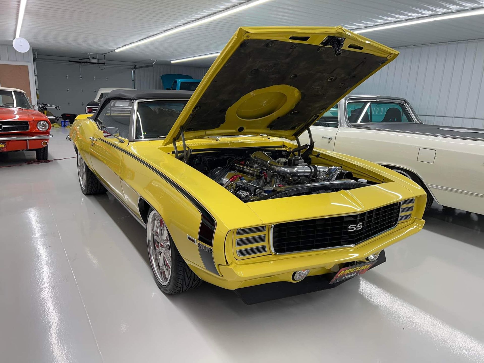 Yellow classic muscle car with hood open in a showroom, parked beside other vintage cars.