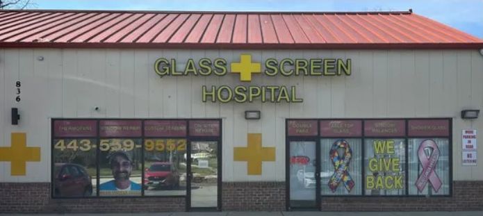 Exterior of the Glass Screen Hospital, featuring a yellow cross logo, shop windows, and a phone number.