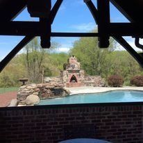 Pool area with a brick structure, stone fireplace, and blue water, seen through a wooden archway.
