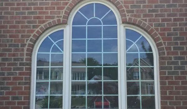 Arched window with white frames, divided by grids, on brick wall; reflections of trees and building in the glass.