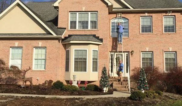 Two people on a ladder decorating a brick house for Christmas.