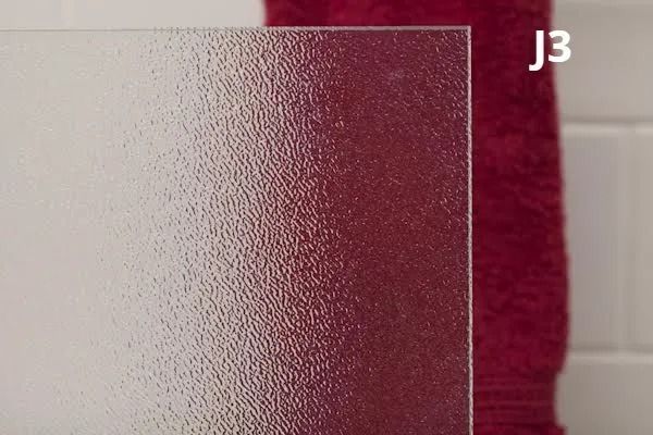 Close-up of a textured glass sheet with a bumpy surface, revealing a distorted view. Red towel and white tiles are blurred in the background.