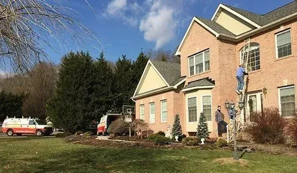 Two-story brick house with workers on a ladder. A van is parked on the lawn. Blue sky, sunny day.