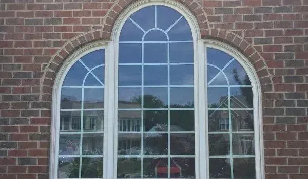 Triple-pane arched window with white frames in red brick wall; sunlight reflects trees and buildings.