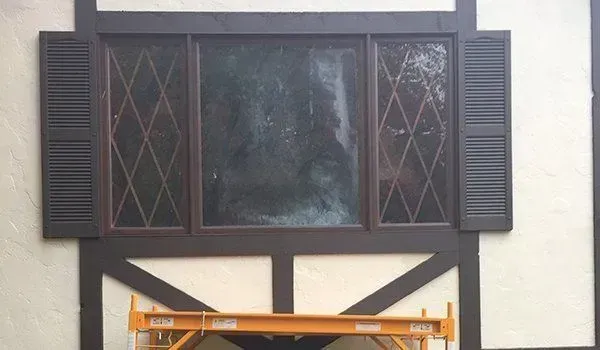 Window with brown diamond-paned glass and shutters, set in a brown and tan building with scaffolding below.