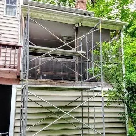Scaffolding erected on the side of a house for repairs; wood siding, a screened porch, and foliage.
