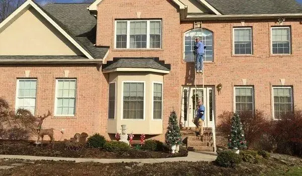 Person on ladder cleaning window of brick house; Christmas decorations visible.