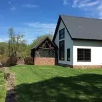 Two-story white house with black trim and a connected wooden structure, on a green lawn. Sunny day.