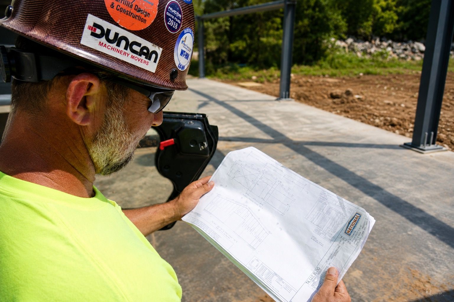 Construction worker in a hard hat reading a plan on a sunny sidewalk, holding a radio