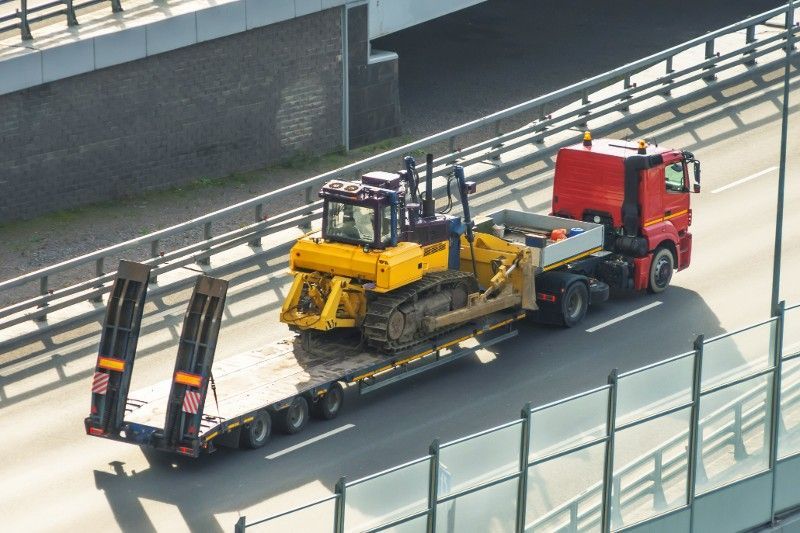 A red truck is carrying a yellow bulldozer on a trailer on a highway.