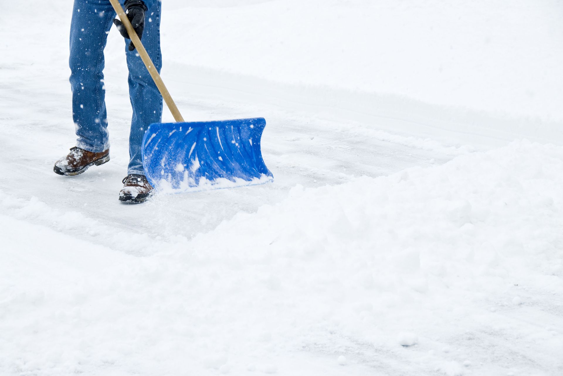 Person shoveling snow with a blue shovel on a snowy surface, wearing blue jeans and work boots.