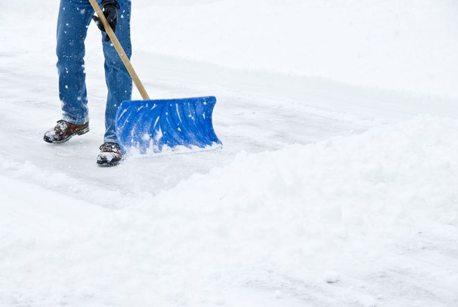 Person shoveling snow with a blue shovel on a snowy surface, wearing blue jeans and work boots.