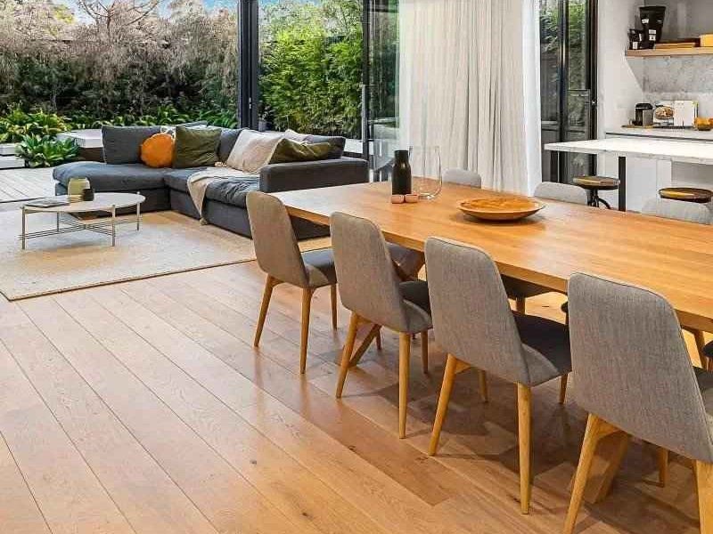Modern dining area with wooden table, gray chairs, gray sofa, and large windows overlooking greenery