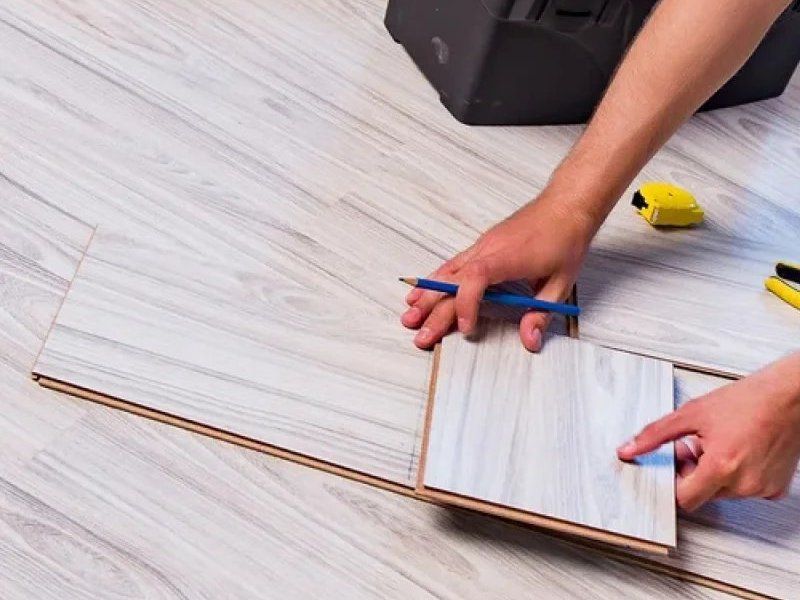 Hands measuring light wood-look flooring with a pencil and tape measure during installation