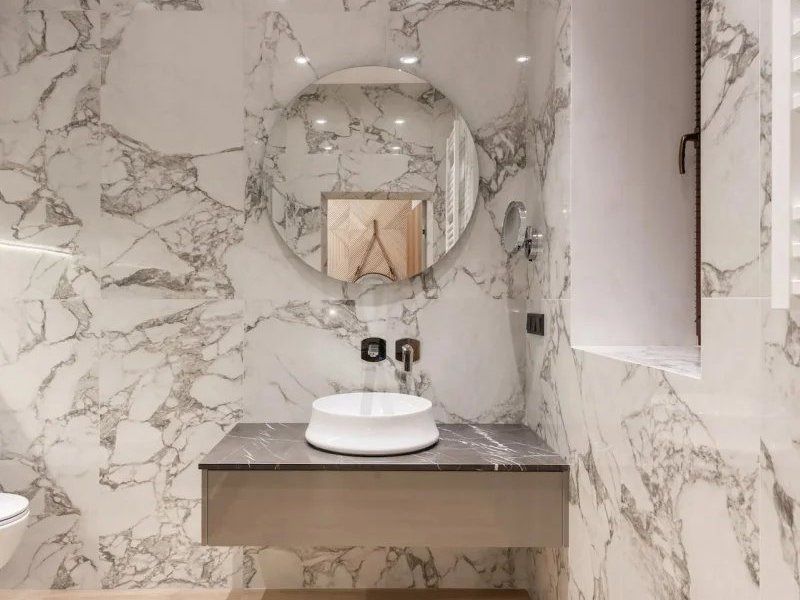 Small bathroom vanity with marble walls, round mirror, vessel sink, and gray countertop.