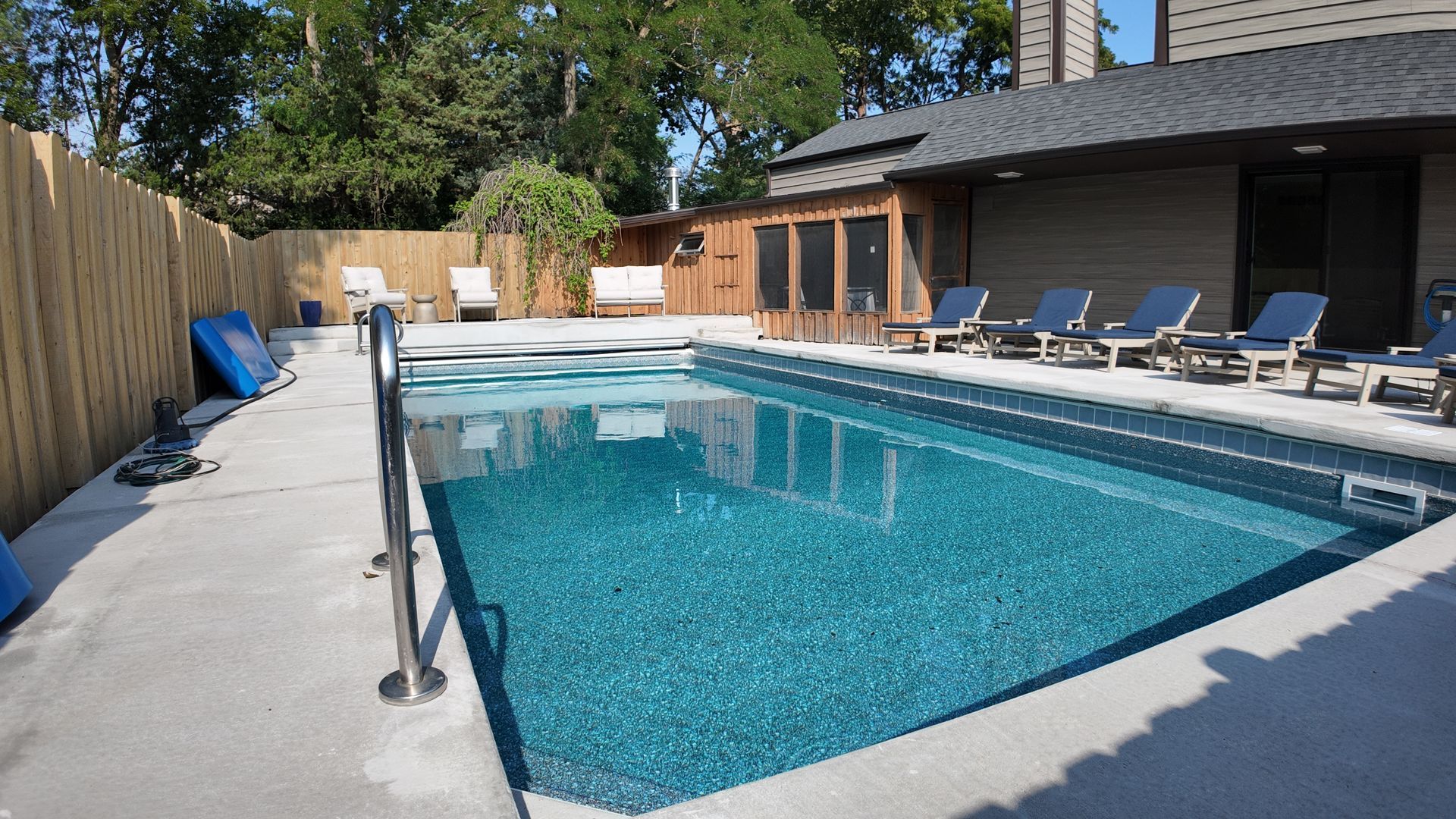 Backyard swimming pool with lounge chairs, wooden fence, and a modern house in the background