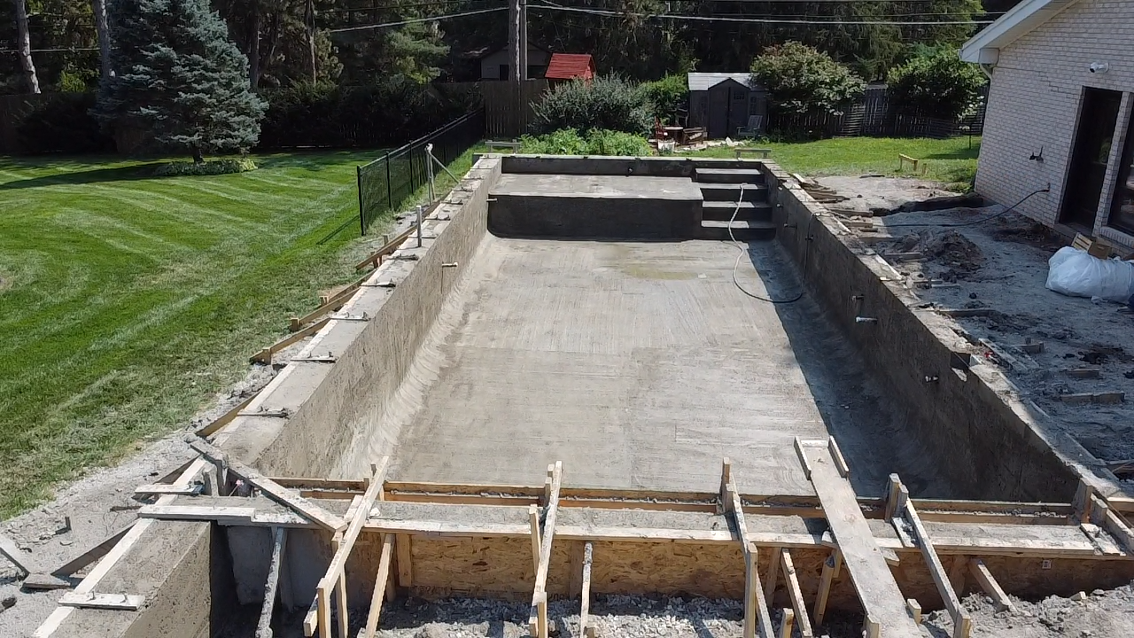 Empty in-ground swimming pool under construction in a backyard, with concrete walls and wooden framing.