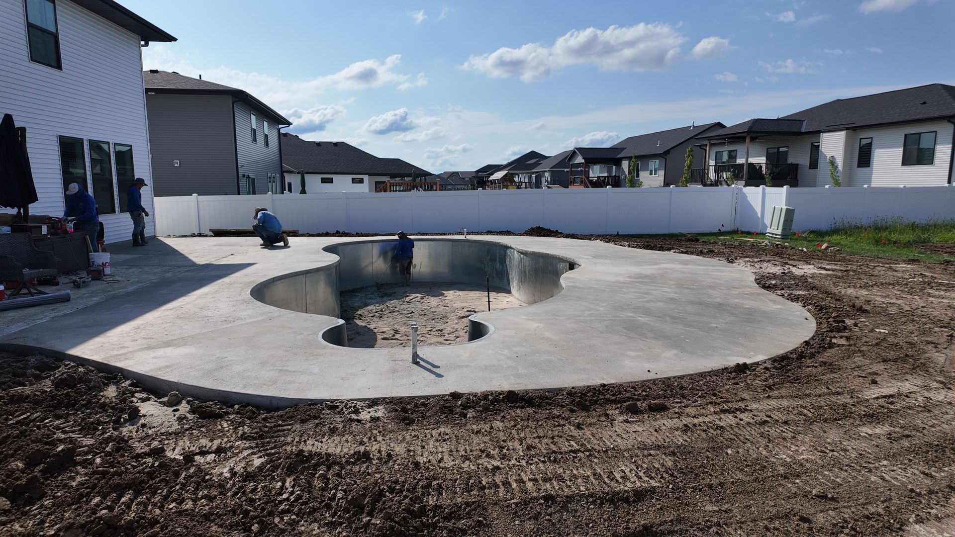 Empty concrete pool under construction in a suburban backyard, surrounded by houses and dirt.
