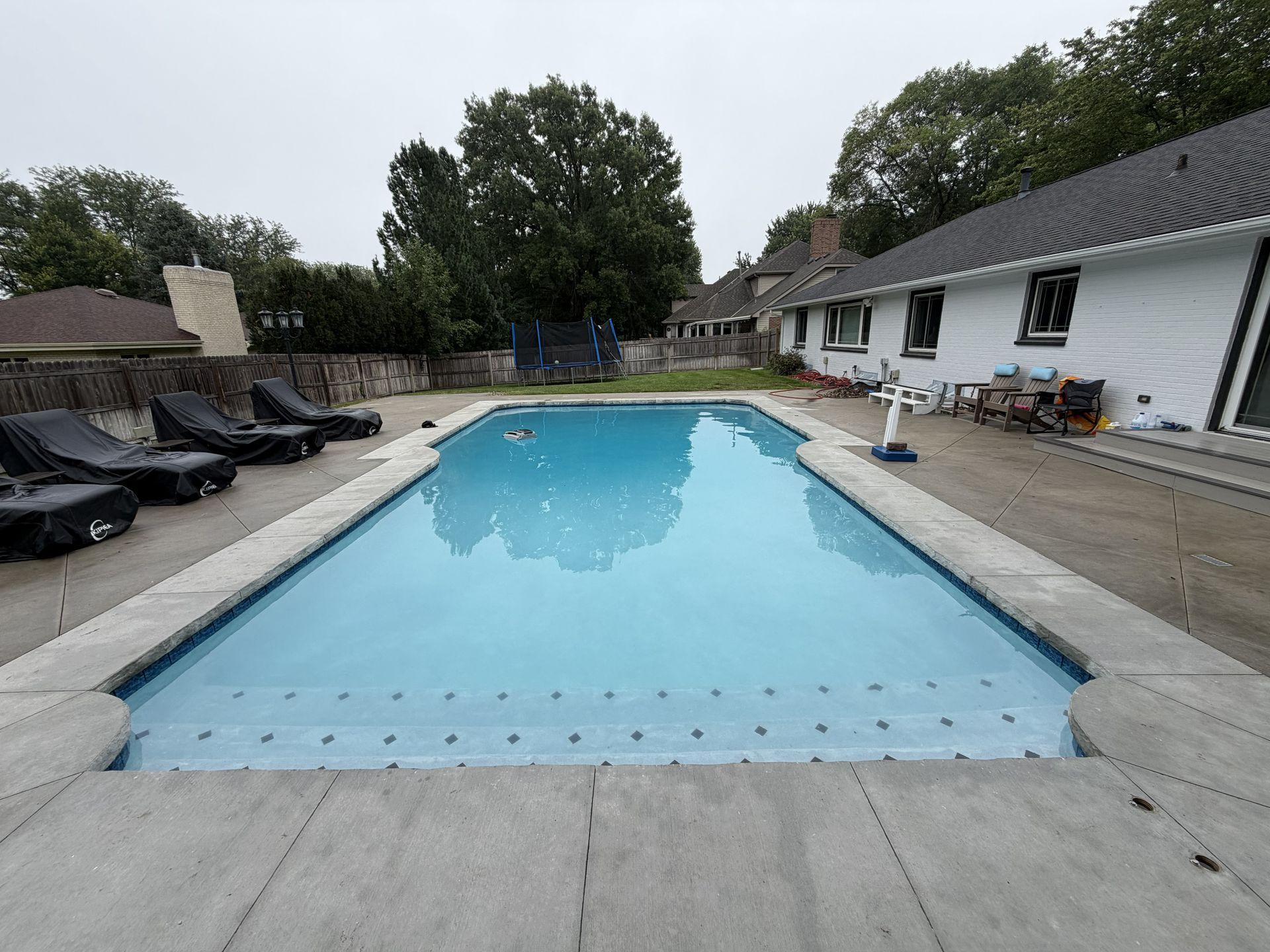 Outdoor swimming pool beside a white house, with a diving board and patio furniture under cloudy skies