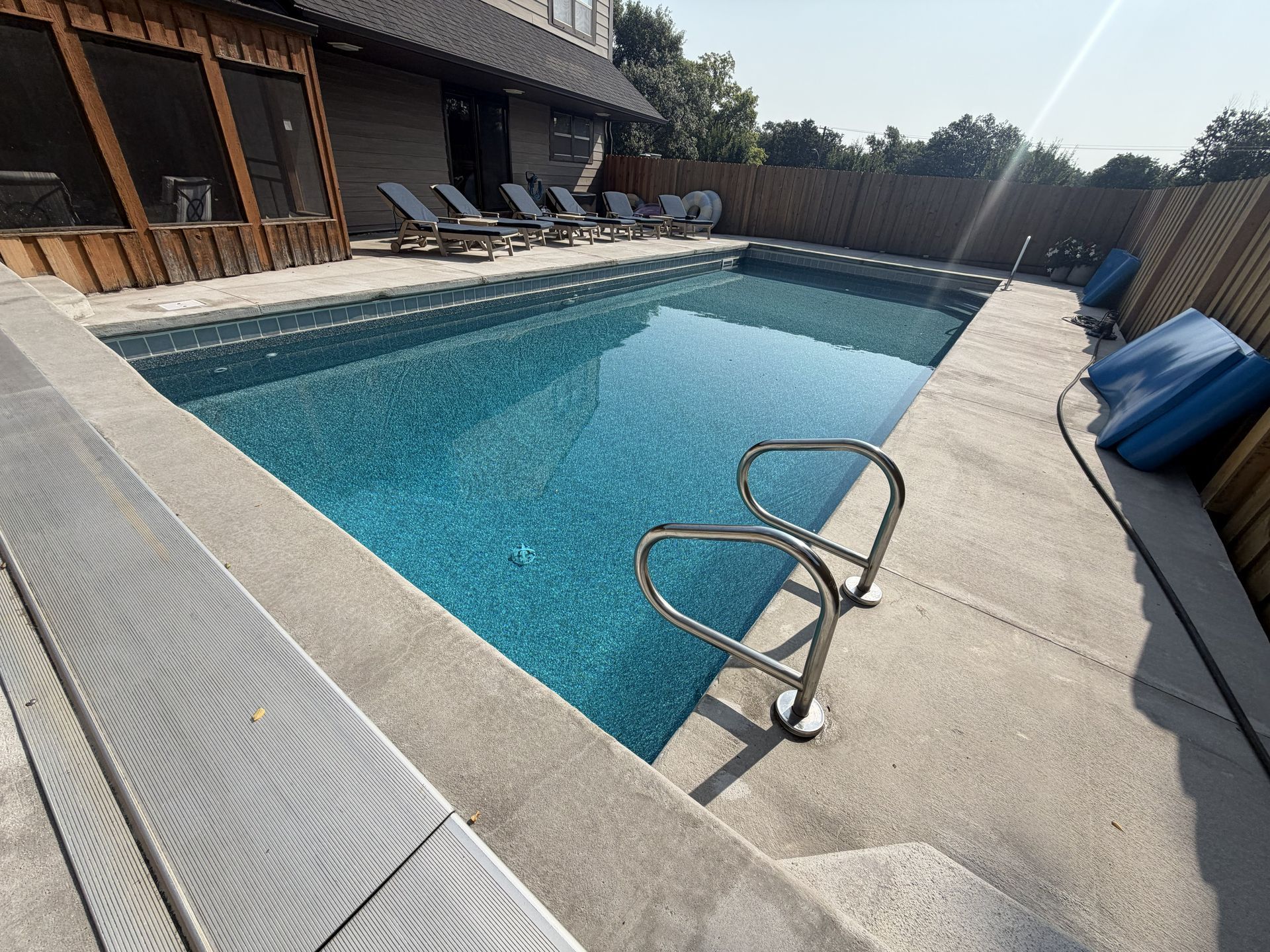 Backyard swimming pool with stone deck, metal ladder, lounge chairs, and blue floating cushions