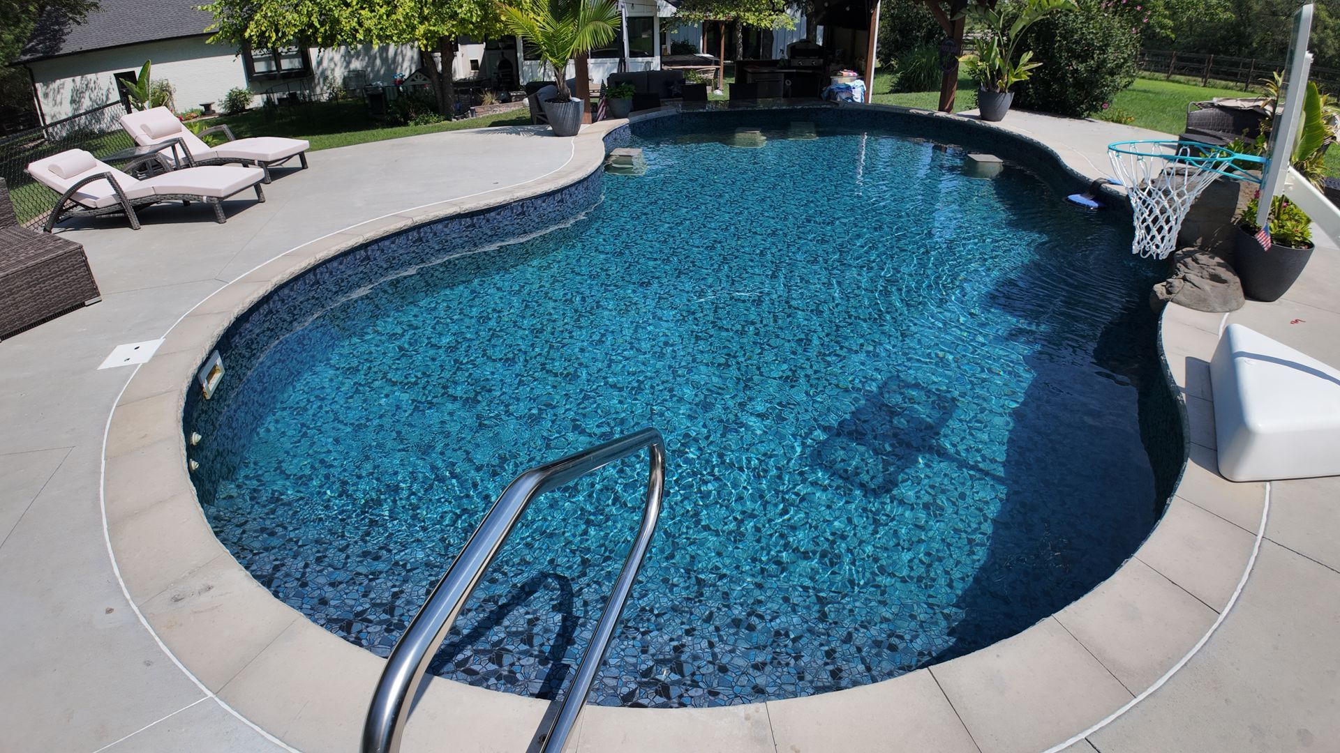 Backyard swimming pool with clear blue water, curved stone deck, and lounge chairs in the background