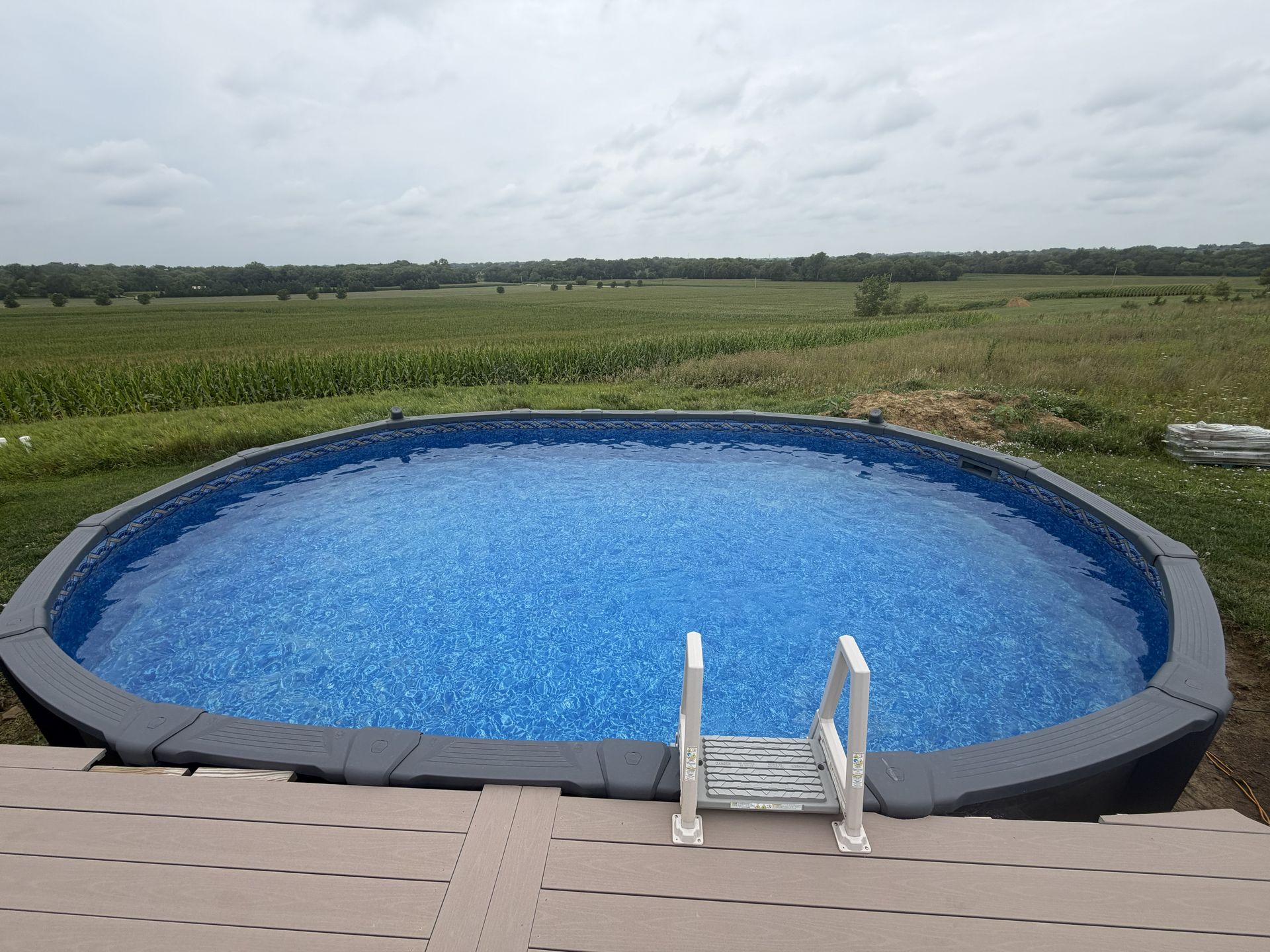 Blue round above-ground pool on a deck overlooking open fields under a cloudy sky
