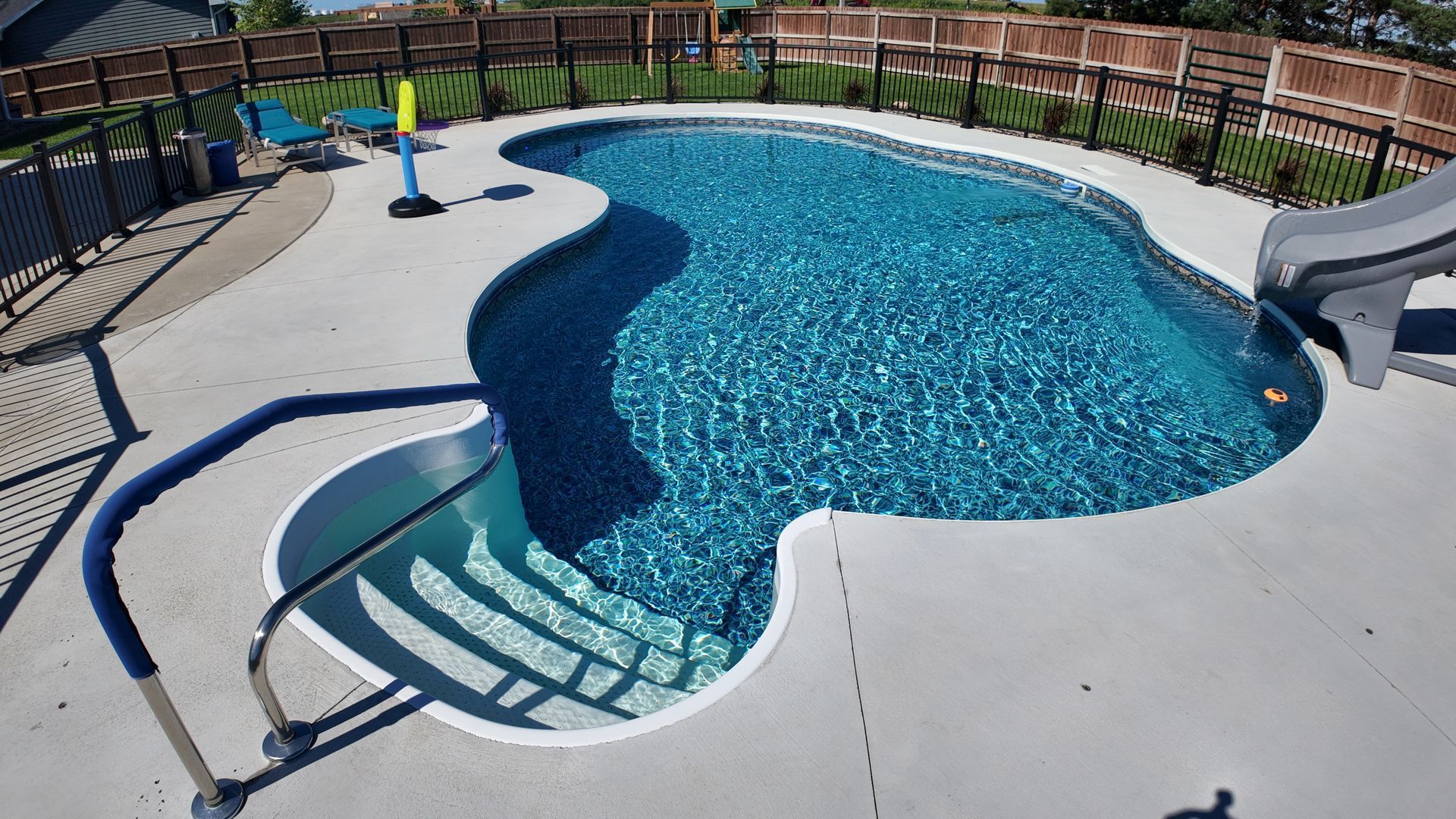 Backyard swimming pool with curved steps, blue water, and a slide on a concrete deck surrounded by a fence