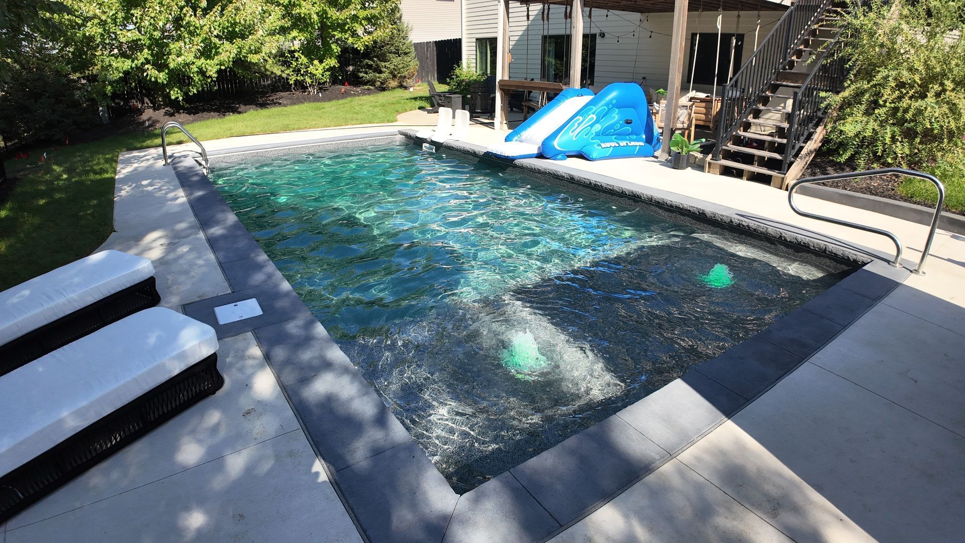 Backyard swimming pool with clear water, lounge chairs, and a blue slide under a shaded patio