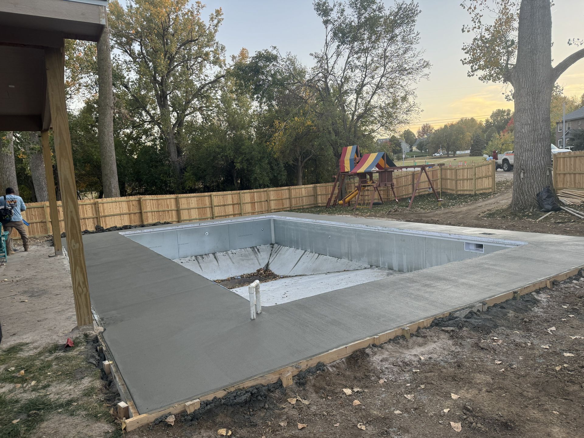 Concrete pool under construction with a wooden fence, trees, and playground equipment in the background