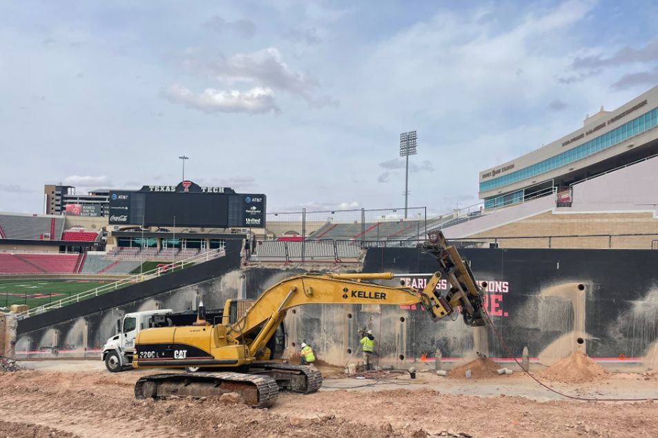 A large yellow excavator is sitting in the dirt in front of a stadium.