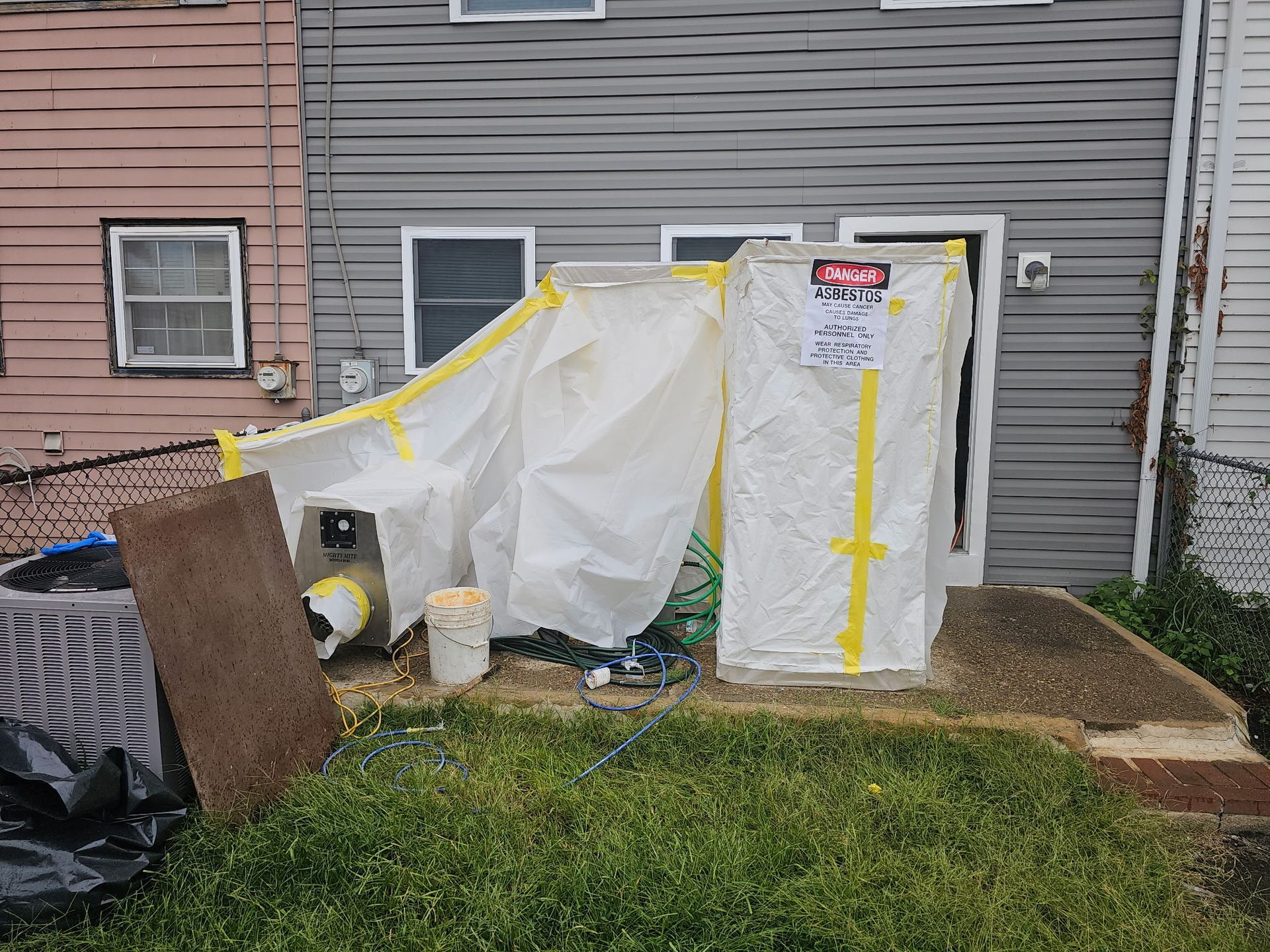 Plastic-covered enclosure with yellow tape beside a house, with a black tarp and wood panel in the yard