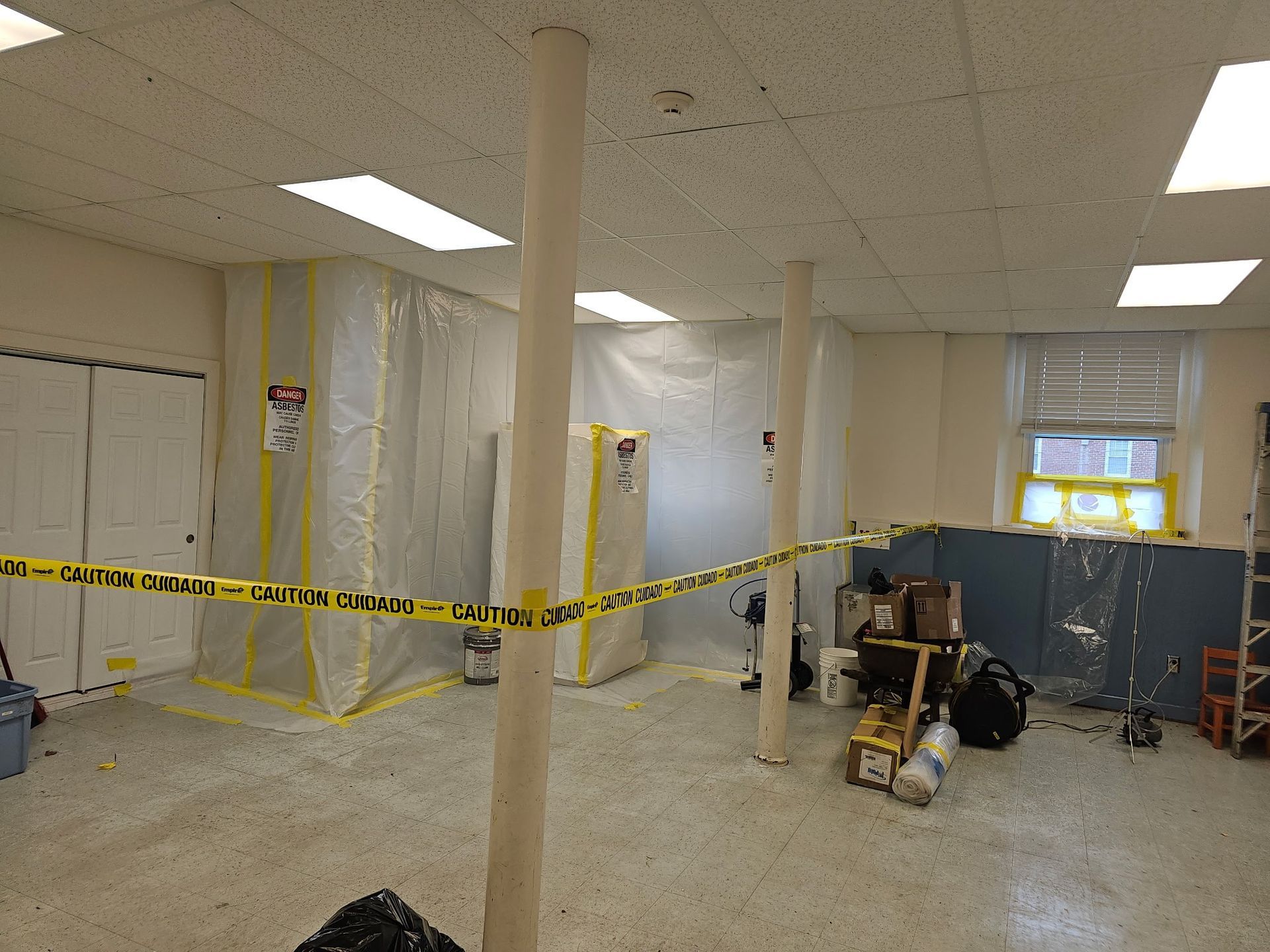 Caution-taped unfinished room with columns, tools, and covered equipment under renovation without ceiling tiles