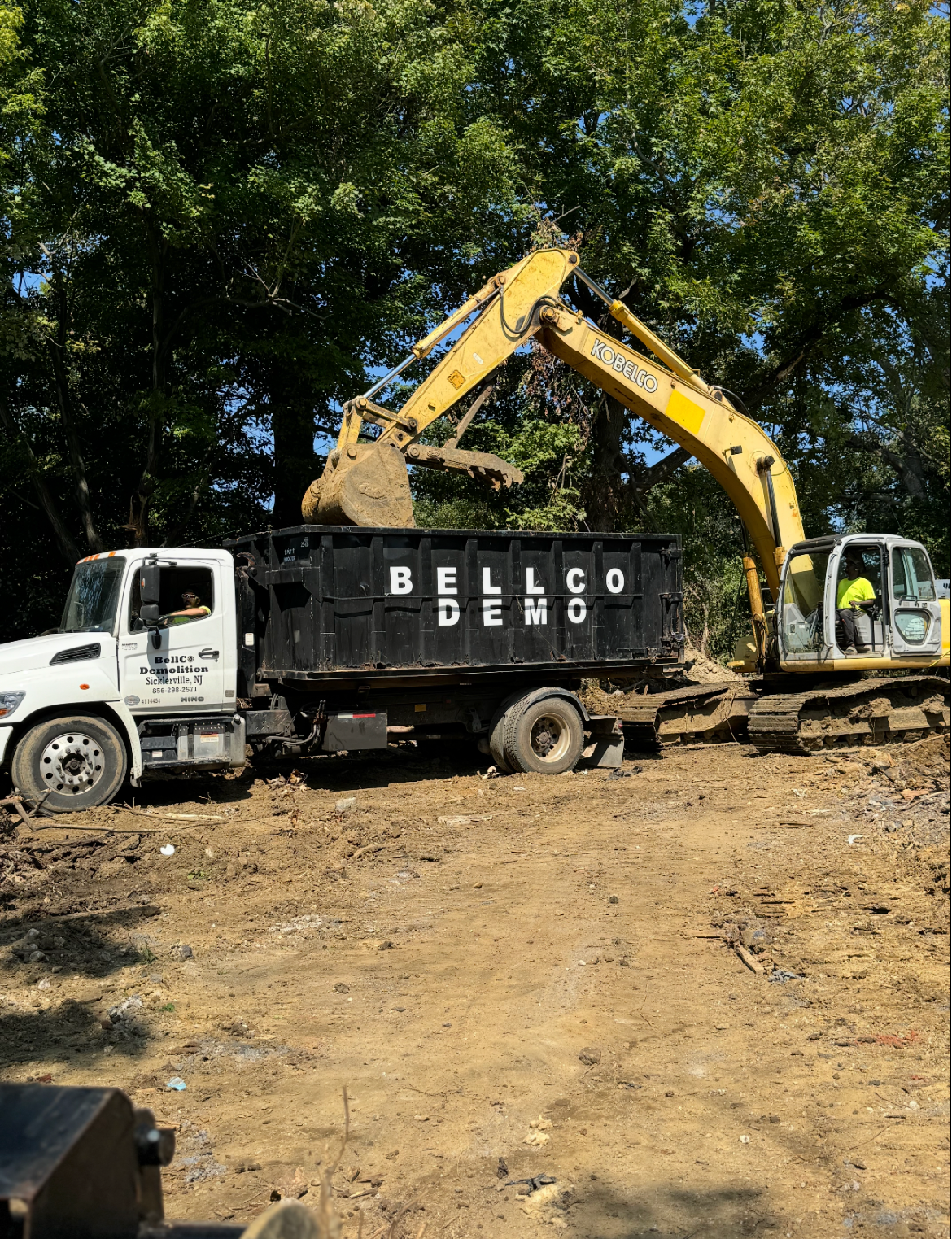Excavator loading debris into a black Bellco dump truck at a construction site under trees