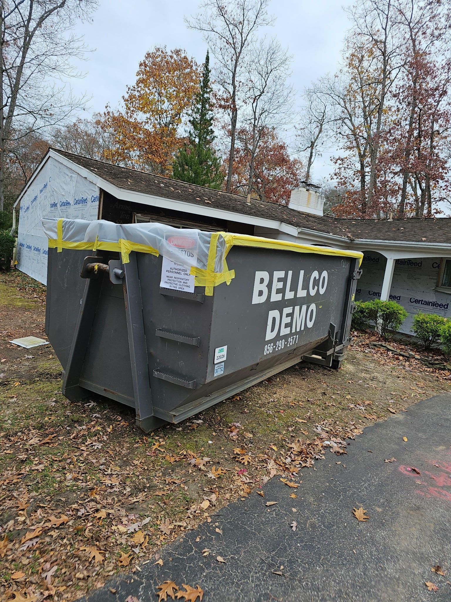Large gray dumpster with yellow tape and a white sign in a wooded roadside area